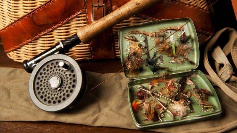 The image displays a collection of fly fishing gear arranged on a wooden surface. At the center is a fly reel attached to a rod, with a weathered handle suggesting regular use. Next to it is an open green metal box containing a variety of colorful fishing flies, meticulously organized in compartments. The flies are crafted with feathers, fur, and threads, designed to mimic different insects. In the background, a woven wicker fishing basket with leather straps adds a rustic touch to the scene.