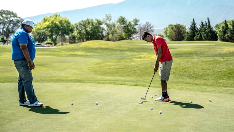 Three golfers are standing on a lush green golf course with a backdrop of dense pine trees and distant mountains under a partly cloudy blue sky. One golfer is mid-swing, while the other two watch, holding their clubs. 