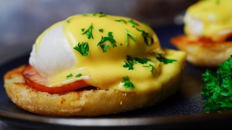 The image shows a close-up of a classic Eggs Benedict dish. A poached egg is placed on top of a slice of ham or Canadian bacon, which sits on a toasted English muffin. The egg is generously covered with creamy hollandaise sauce and garnished with fresh parsley.