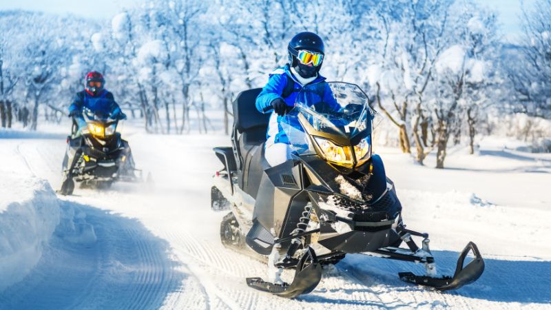 Two people riding snowmobiles through a snowy landscape.