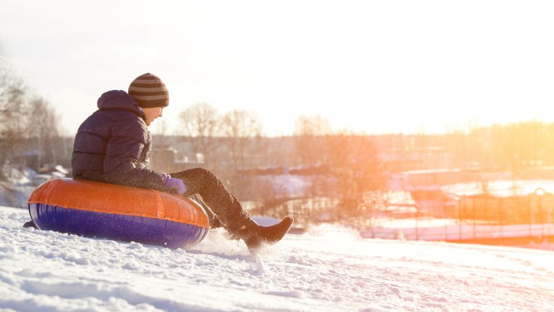 A person in a winter jacket and beanie is sitting on an orange and blue inflatable snow tube, preparing to slide down a snowy hill. The scene is bathed in warm sunlight, with a snowy landscape and leafless trees in the background, evoking a peaceful winter day outdoors.