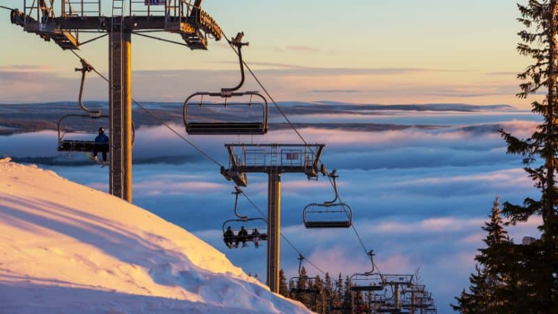 A scenic view of a ski lift at sunset, with chairs suspended over a snow-covered mountain. A layer of clouds and fog settles below, creating a serene and mystical atmosphere.