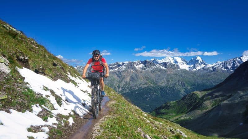 A mountain biker rides along a narrow trail with a stunning view of green hills, snowy patches, and distant mountain peaks under a bright blue sky.