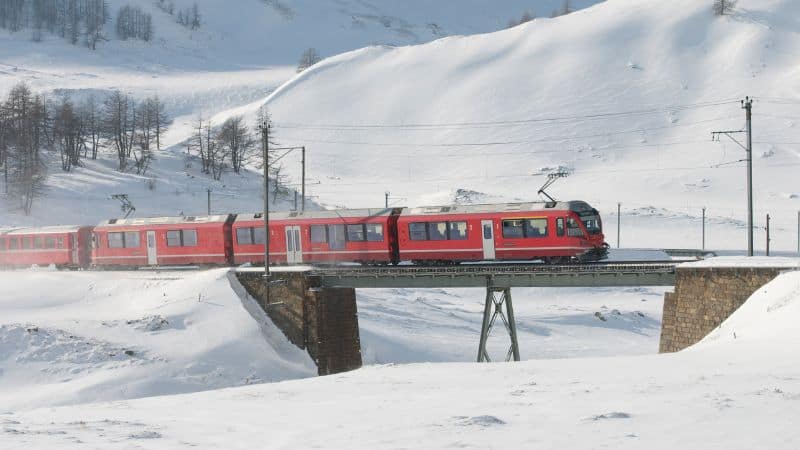 A red train travels across a bridge over a snowy landscape, with snow-capped mountains and a bright, clear sky in the background.
