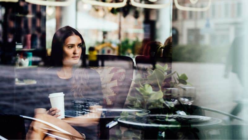 A woman holding a coffee cup while sitting in a trendy coffee shop, viewed through a window with reflections of outdoor scenery and plants in the foreground.