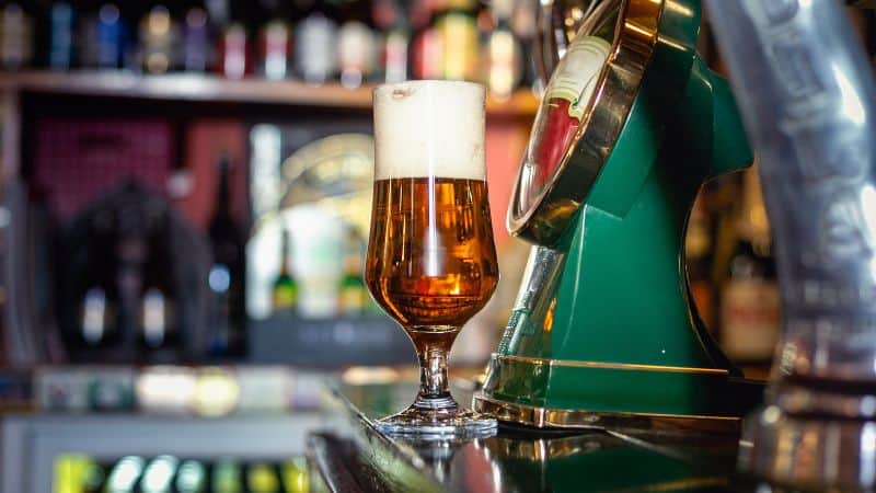 A crisp glass of beer sitting on a bar counter, with bottles and bar decor softly blurred in the background.