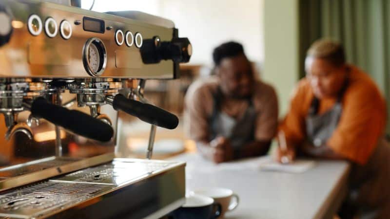 A professional espresso machine on a counter in a coffee shop, with two baristas sitting at a nearby table in discussion.