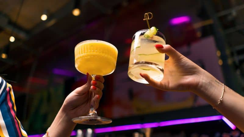 Two hands holding different cocktails in an illuminated bar, with vibrant colors and a festive atmosphere.