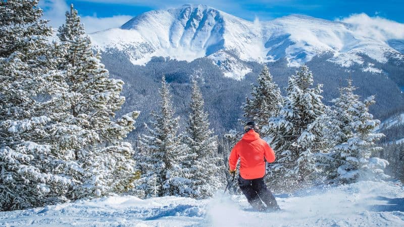 A skier dressed in a bright red jacket skiing downhill on a snowy mountain surrounded by pine trees, with a stunning mountain peak in the background.