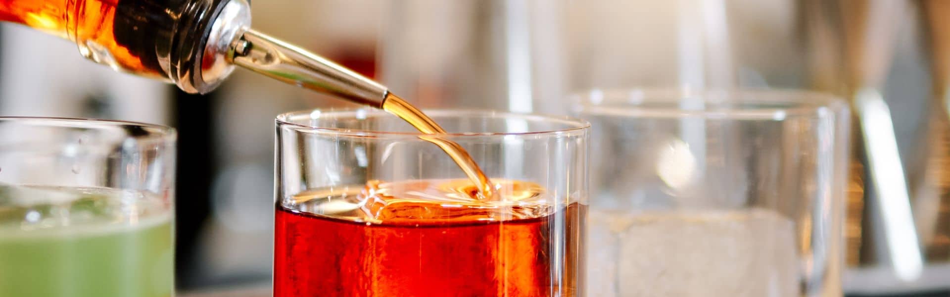 A close-up shot of amber-colored liquid being poured into a glass with a stainless steel pour spout, surrounded by other drinks in the background.