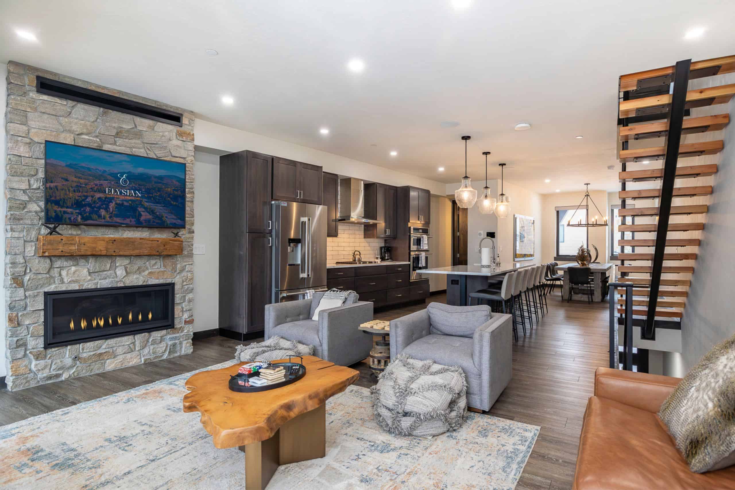 Modern open-concept living space featuring a stone fireplace with a mounted TV, a sleek kitchen with dark cabinetry and stainless steel appliances, a large island with bar seating, and a floating wood staircase with black metal accents. The room is furnished with cozy gray armchairs, a live-edge wooden coffee table, and a leather sofa, creating a warm and inviting atmosphere. A dining area with a statement chandelier is visible in the background.