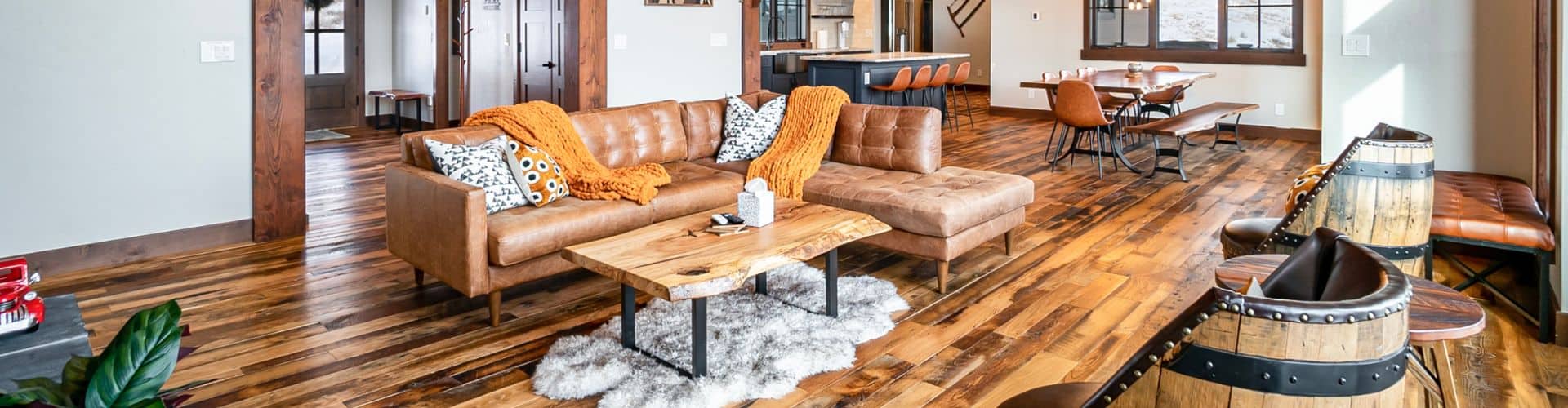 A spacious, open-concept living area with rich wood flooring, a plush leather sectional adorned with vibrant throw blankets, and a rustic wood coffee table. The room flows into a stylish kitchen and dining area, offering a perfect blend of modern and rustic aesthetics.