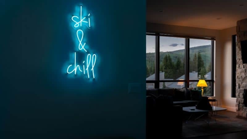 A neon blue &ldquo;ski & chill&rdquo; sign glows against a dark blue wall in a dimly lit lounge area. Large windows reveal a serene mountain view in the background, setting a cozy apr&egrave;s-ski ambiance.