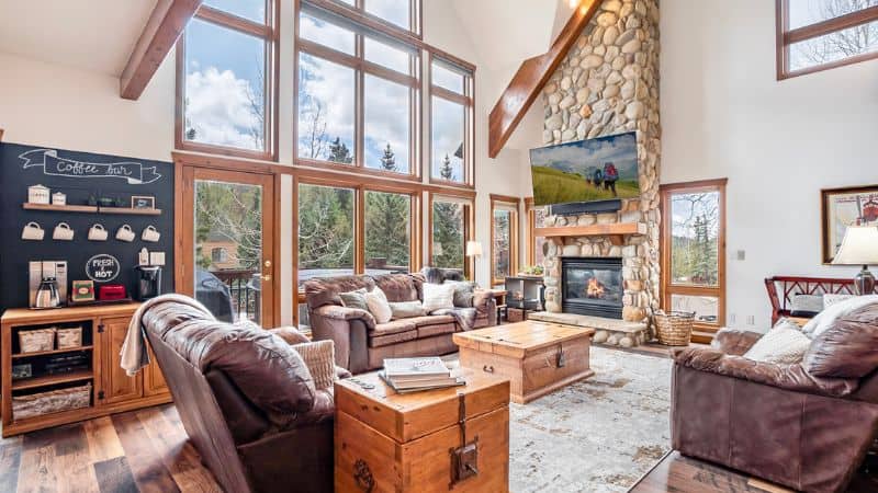 A rustic-modern living room with floor-to-ceiling windows, a towering stone fireplace, and a cozy leather furniture set. A built-in coffee bar adds charm, making the space a warm and welcoming retreat.