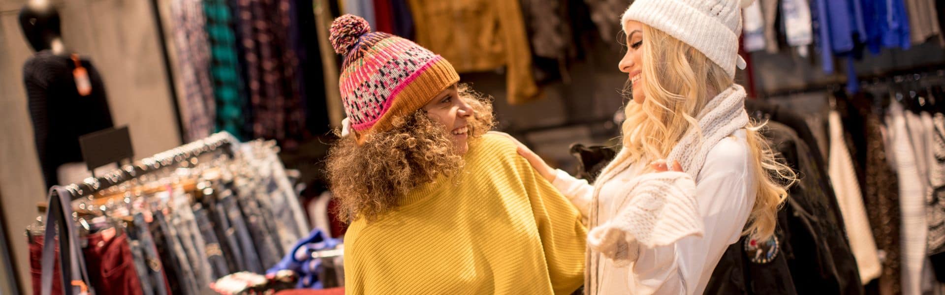Two people shopping in a clothing store. One person wears a bright yellow sweater and a colorful knit hat with a pom-pom, while the other wears a white winter outfit with a white beanie. They appear to be smiling at each other with clothing racks visible in the background.