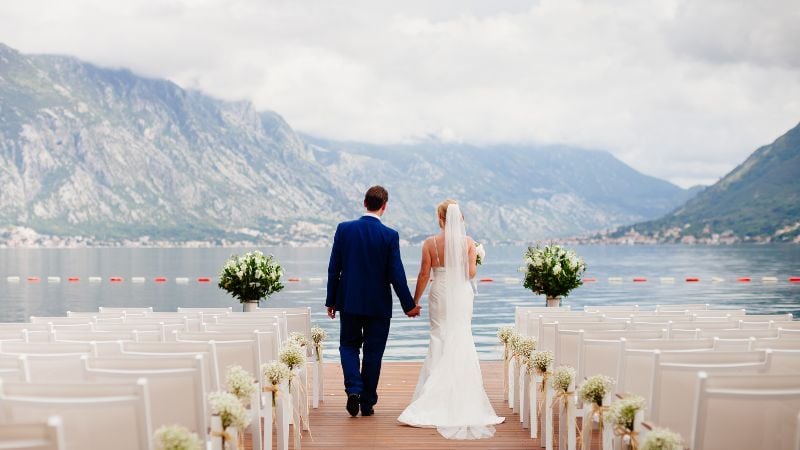 Bride and groom walking hand in hand down a dock surrounded by water and mountains.