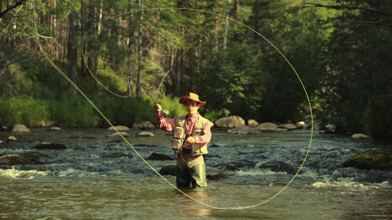Person fly fishing in a shallow river surrounded by green trees.