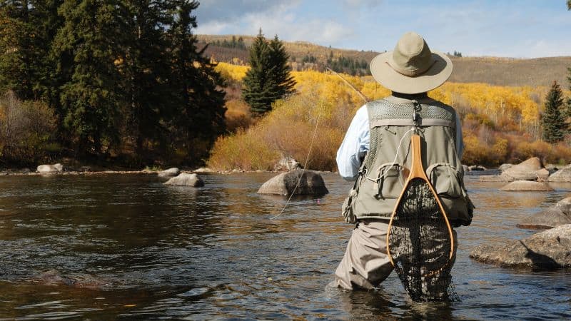 Person fly fishing in a river with autumn trees in the background.