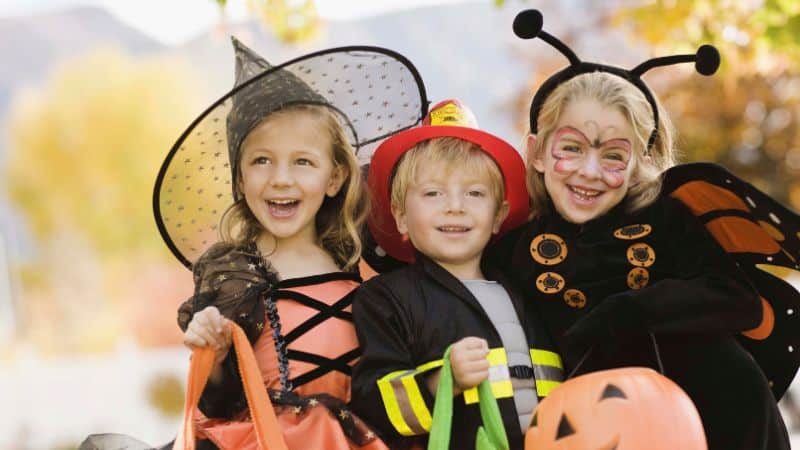 Three children in Halloween costumes smiling and holding treat bags.