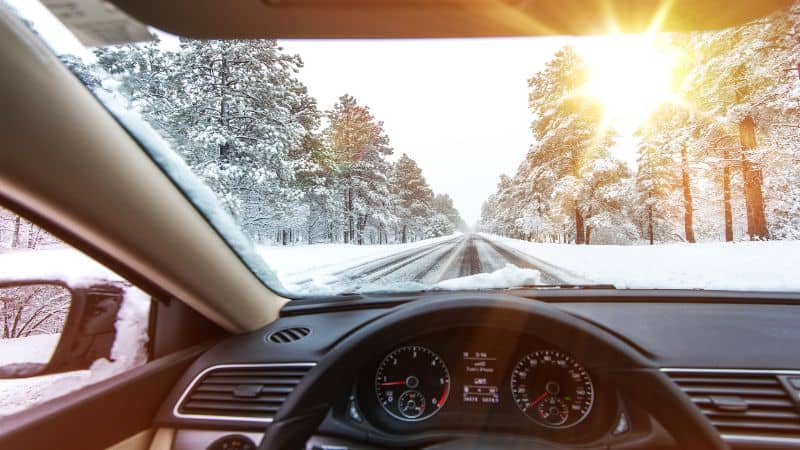 View through a car windshield of a snowy road at sunrise.
