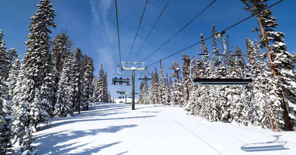 Chairlift carrying skiers over a snowy slope lined with pine trees.