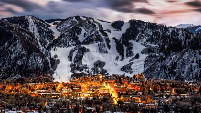 Snow-covered mountain town glowing with lights at dusk beneath ski slopes.