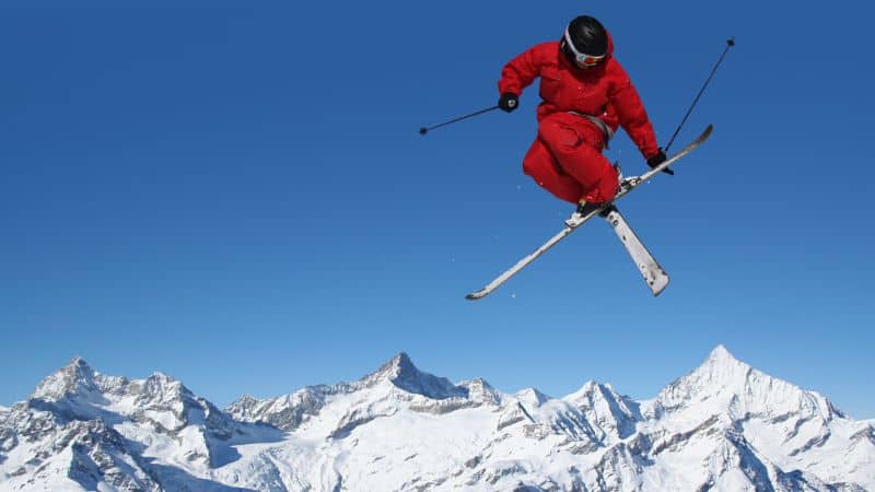 Skier in red outfit jumping high above snowy mountain peaks.