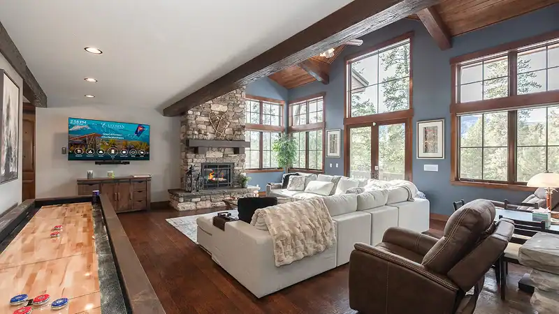 A spacious, rustic-style living room with a large stone fireplace, a sectional sofa, a shuffleboard table, and large windows looking out at the trees.